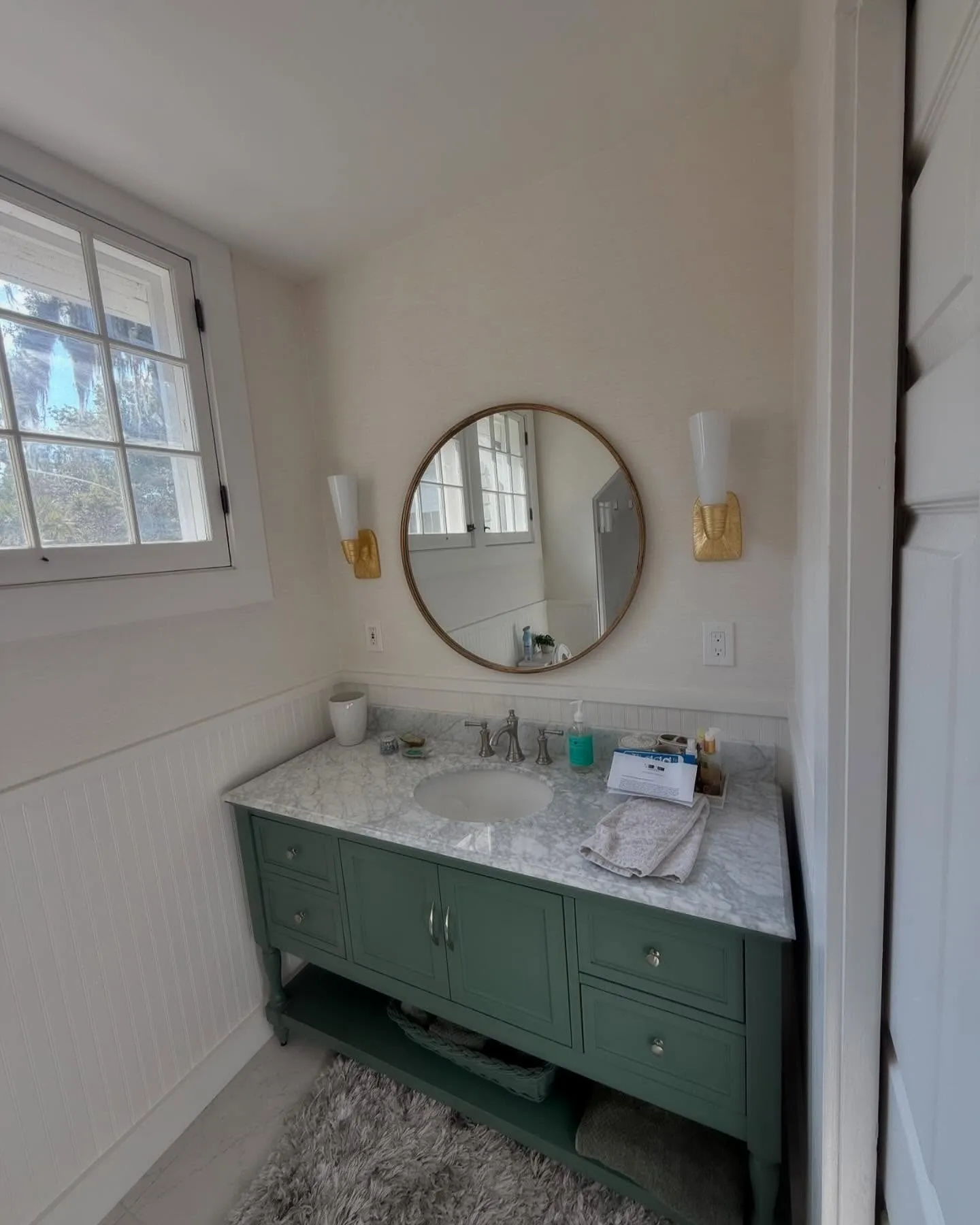 A finished bathroom with a green vanity, round mirror, and a pair of brass wall sconces